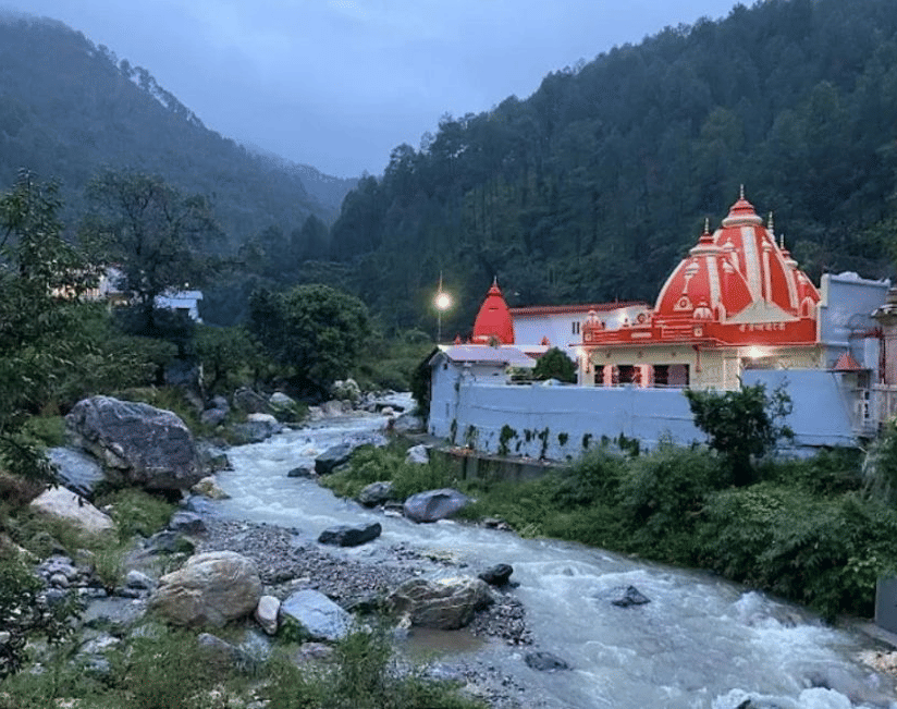  Kainchi Dham temple with a distinctive orange and white shikhara is illuminated beside a rocky river flowing through a densely forested mountain valley at dusk or dawn.