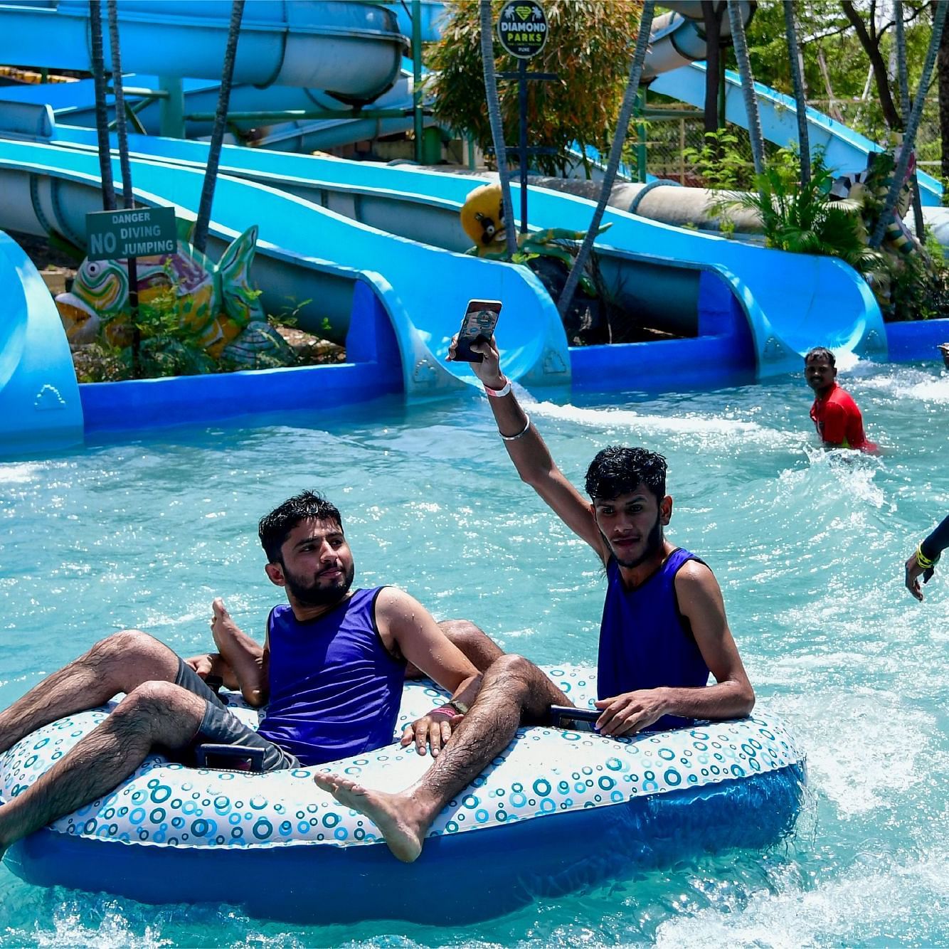 Two people riding inflatable tube through the wave pool at Diamond Parks, Pune, with splashing water and blue water slides visible in the background.