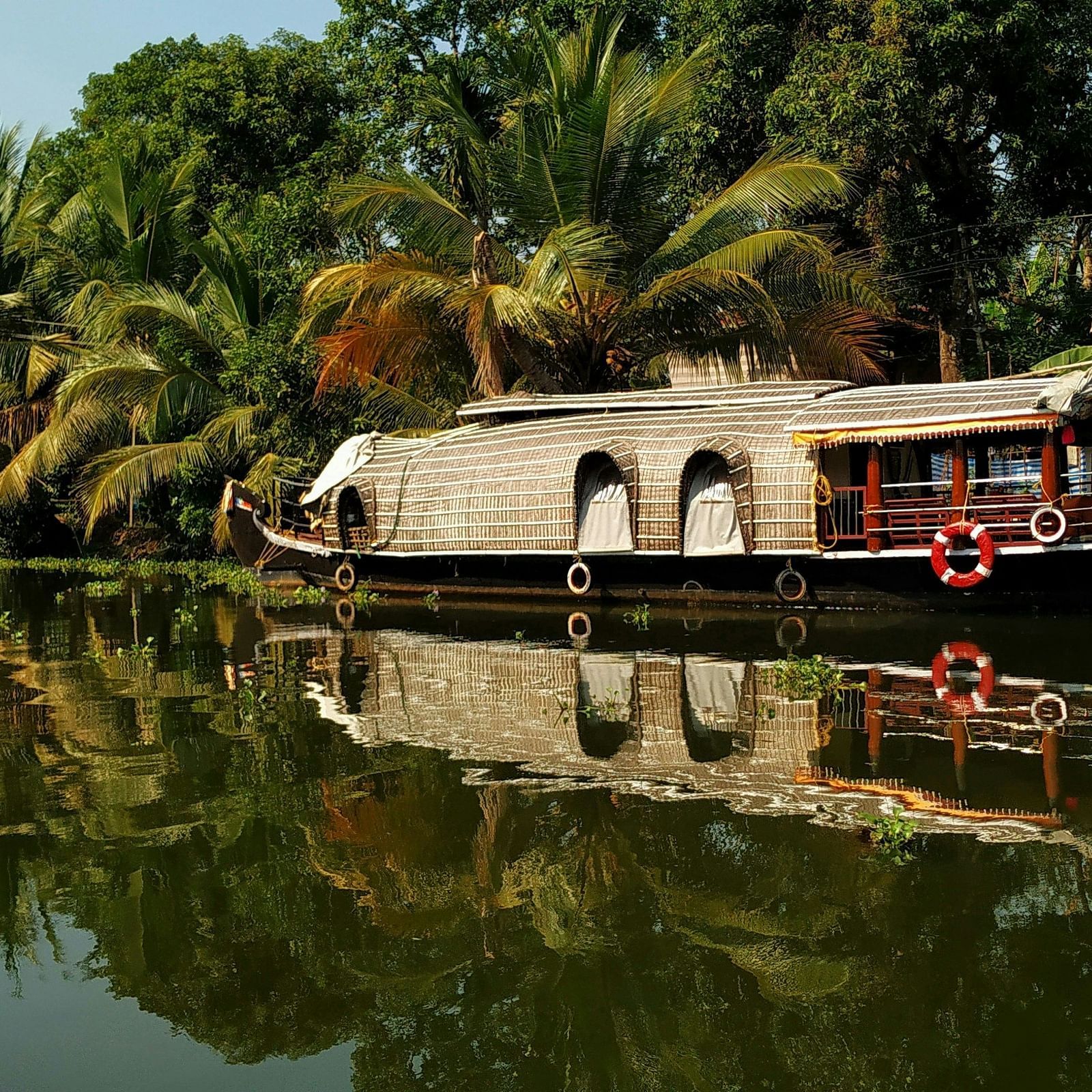 A traditional houseboat with a thatched roof is floating on the calm backwaters, flanked by lush greenery and palm trees reflecting in the water.