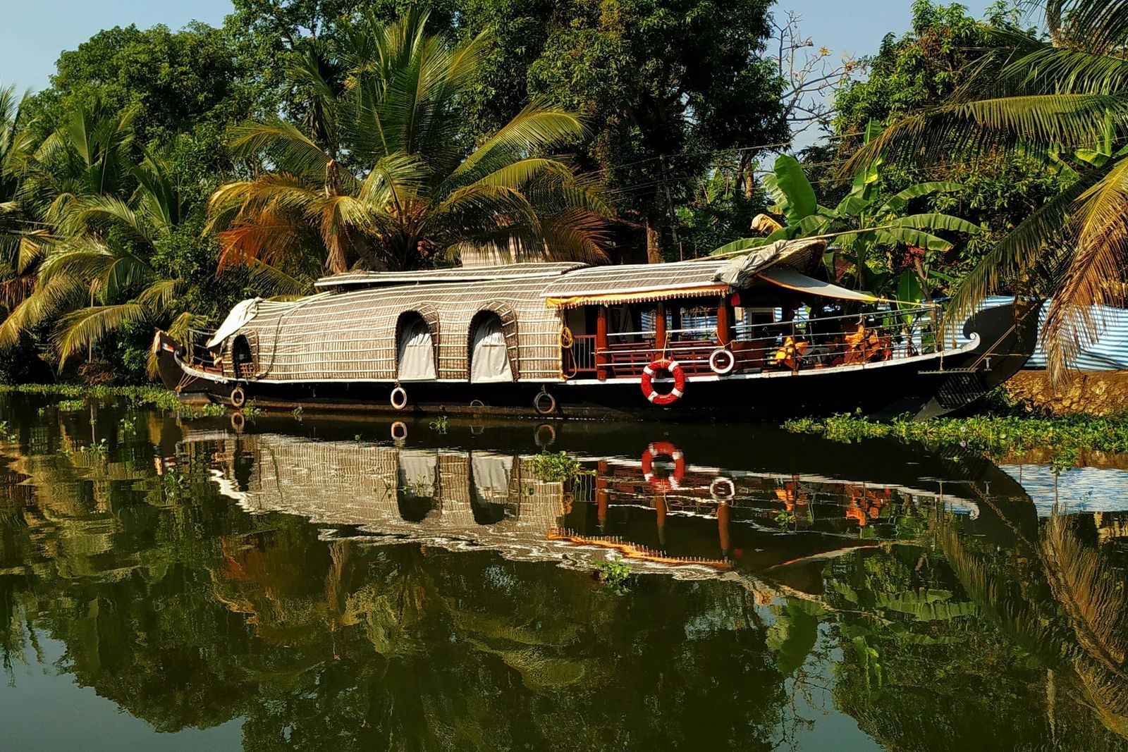 A traditional houseboat with a thatched roof is floating on the calm backwaters, flanked by lush greenery and palm trees reflecting in the water.