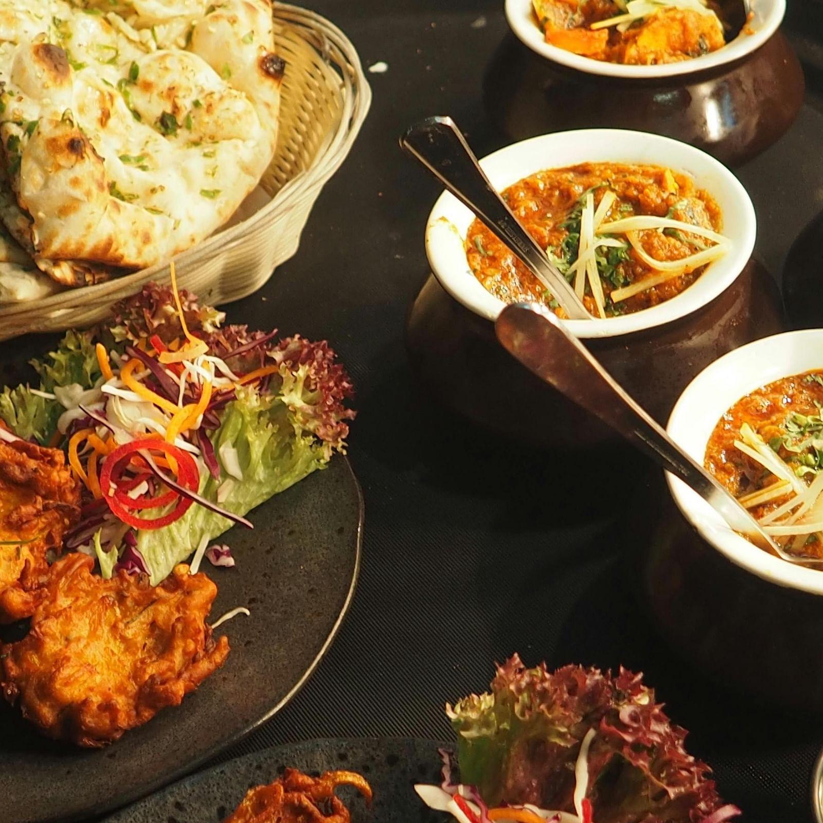 A spread of Indian dishes, including naan, pakora, and curries, is presented on a dark surface.