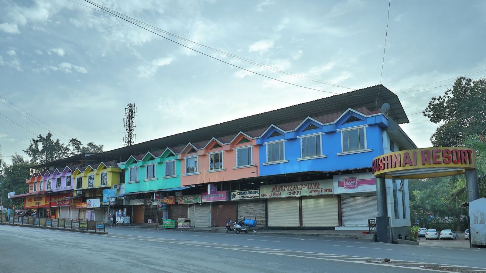 The Krushnai Resort sign marks a long building with colourful facade lining a quiet road under a cloudy sky.