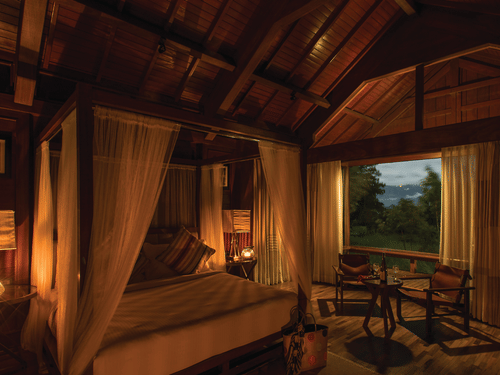 A room at The Serai Bandipur lit up at night with a 4 poster bed,  chairs, and large windows