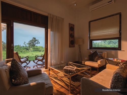 Sun illuminating the interiors of a room at The Serai Bandipur featuring a sofa set and a patio seen through the doorway with a couple of chairs facing the trees outside