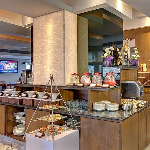 Buffet area at HHI Select, Bangalore featuring a variety of food displays, wooden counters, stacked plates, a TV screen, and modern interior decor. 