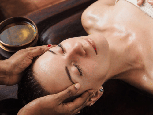 A woman lying on her back with her eyes closed receiving an Ayurvedic head and face massage with oil