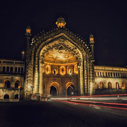A historic, grand arched gateway of the Rumi Darwaza in Lucknow is illuminated at night, with light trails from cars on the street in front of it.