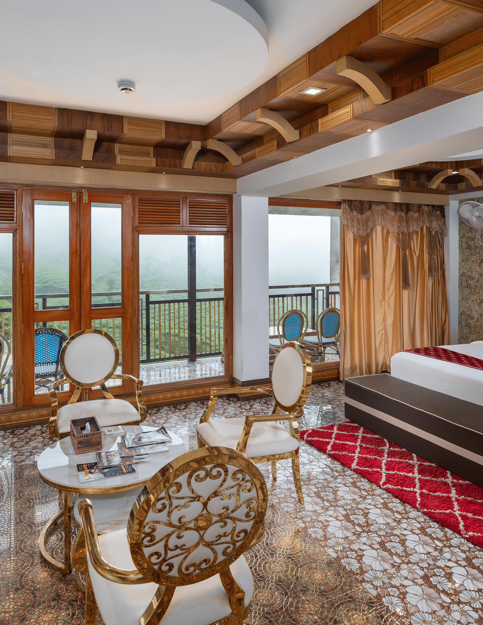 The expansive bedroom of the Presidential Suite at Parakkat Nature Hotel and Resorts, Munnar, featuring a large bed, decorative wooden-beam ceilings, a seating area with a table, and glass doors opening to a scenic view.