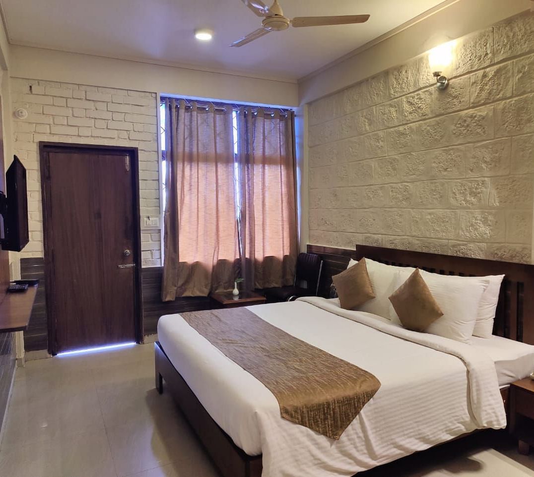 Bedroom with a huge bed, bedside table, windows with curtains, wooden door, and a TV | Standard Room | Woodstock Resorts, Coorg