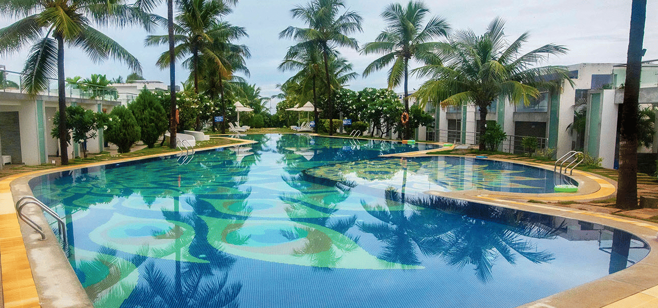 Long swimming pool bordered by coconut trees and cabanas under clear blue skies - Grande Bay Resort & Spa, Mamallapuram