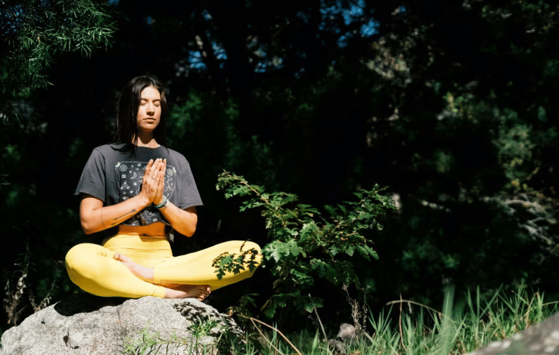 A woman with dark hair meditates peacefully in a lotus position on a rock, wearing pants and a t-shirt, surrounded by nature.