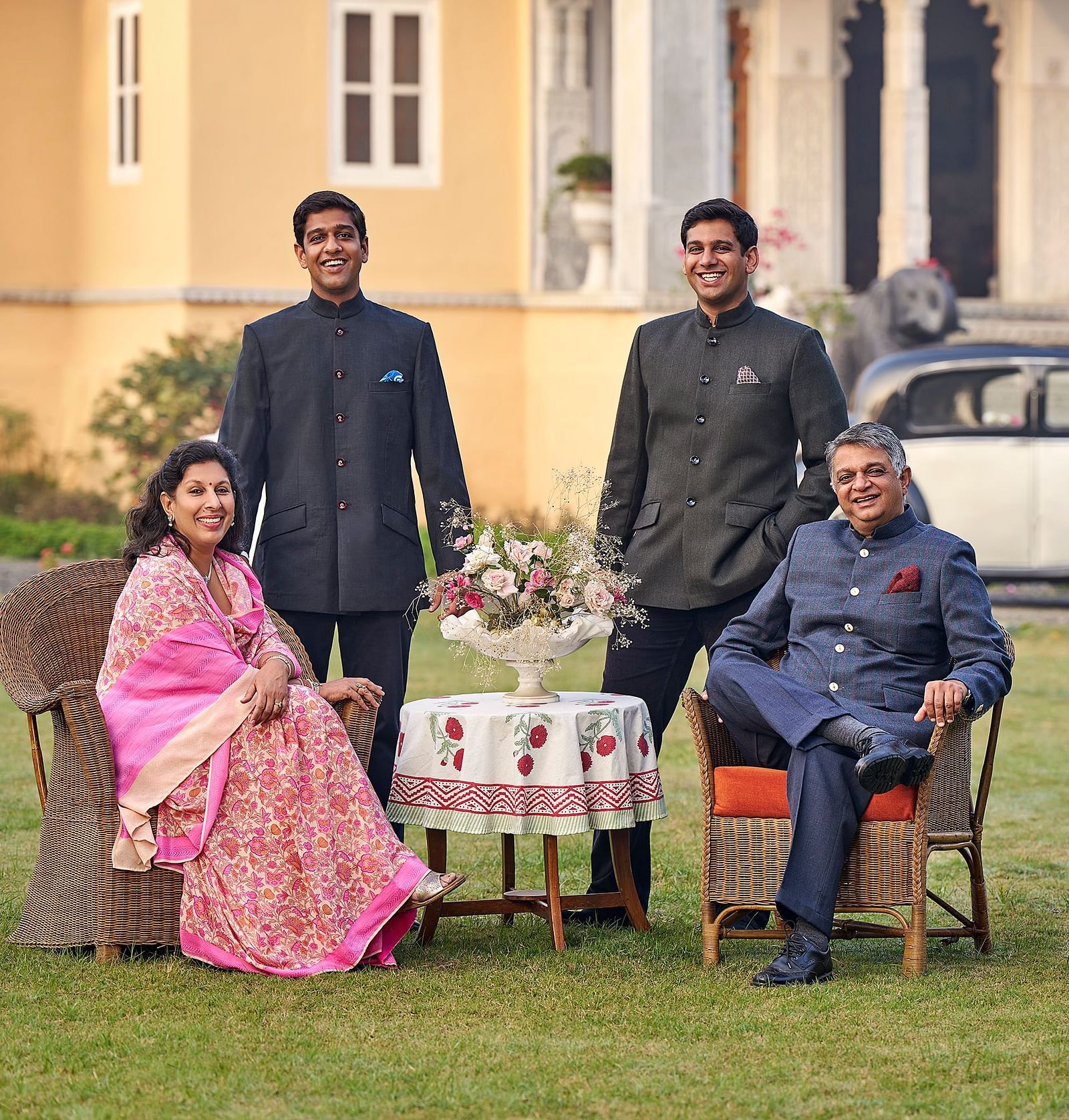 a family portrait of the dev shree family with the parents sitting on the chair while the children stand beside them in a garden