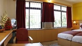 A bedroom with twin beds, a bedside table with a lamp, a chair, a desk, a console table, a mirror and large windows at Hotel Mamallaa Heritage, Mahabalipuram.