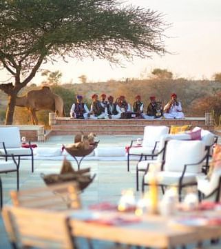 An image of outdoor seating area with seating arrangement placed in front of a podium where a group of men are sitting wearing traditional Rajasthani outfit - MANVAR Resort & Desert Camp