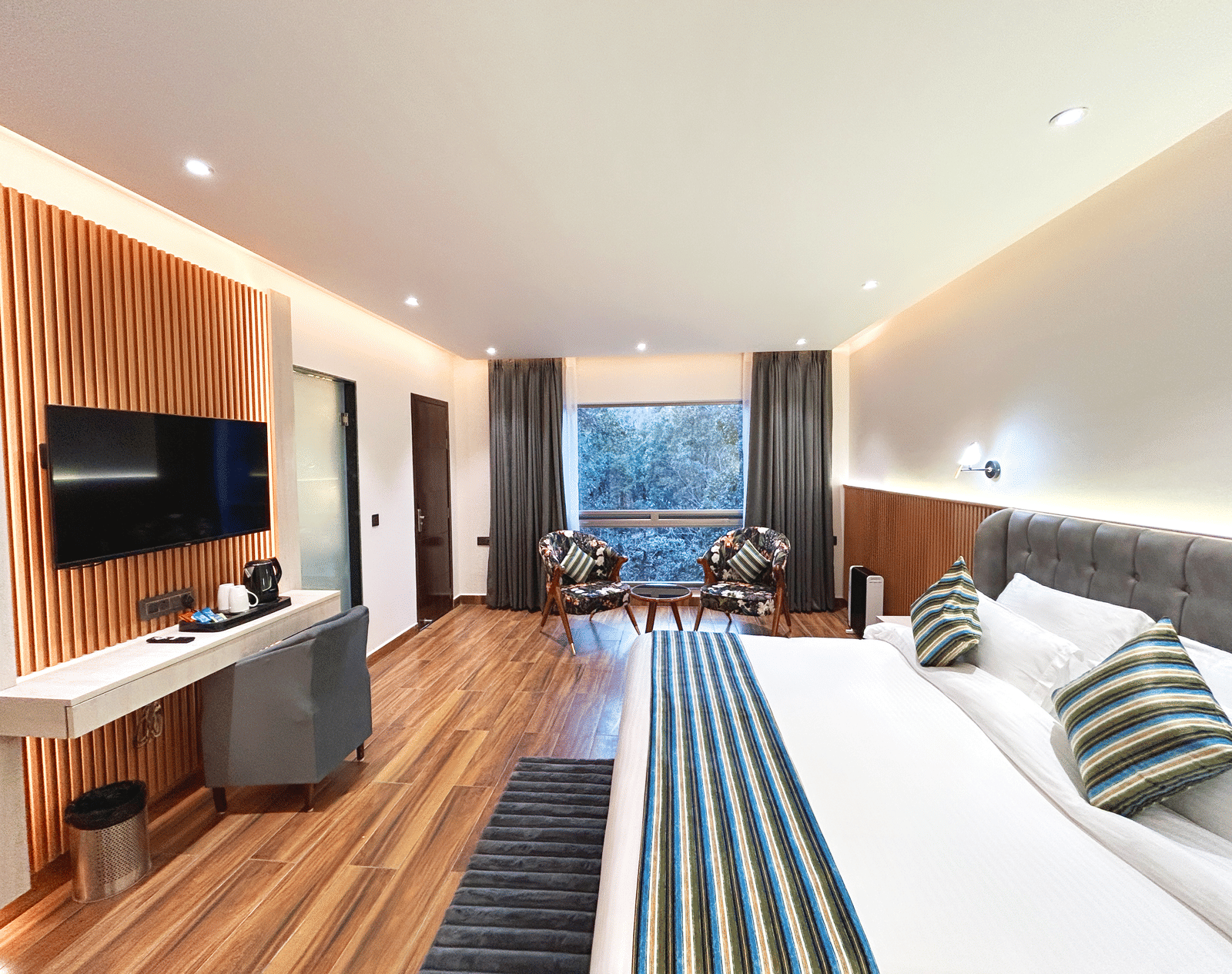 An image of the premium room at DLS The Emerald, Nainital featuring a bed, a flat screen TV, chairs and table and a window overlooking the view