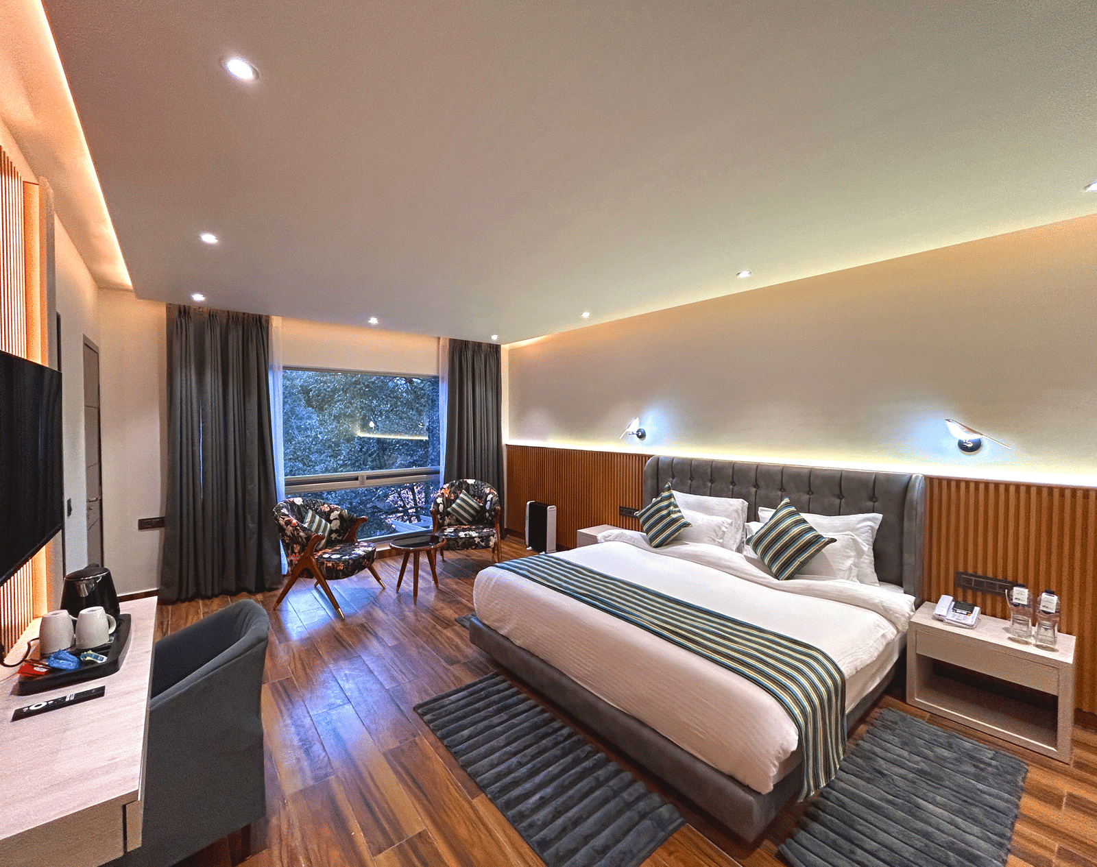 The premium room of DLS The Emerald, Nainital with a bed, bed side table, chairs, coffee table, a window overlooking the view, with warm lighting