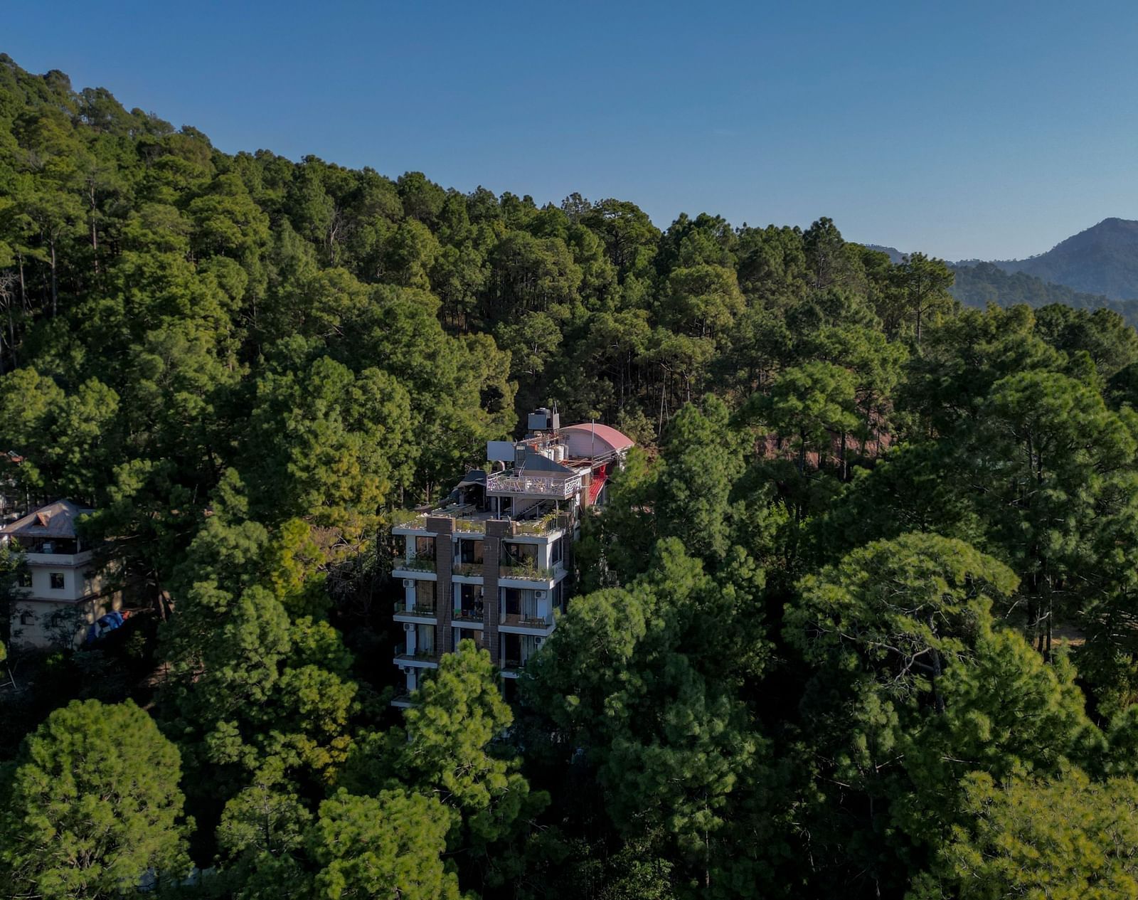 A drone shot taken during the day of DLS Whispering Winds, Kasauli, surrounded by lush greenery and the backdrop of a clear sky.
