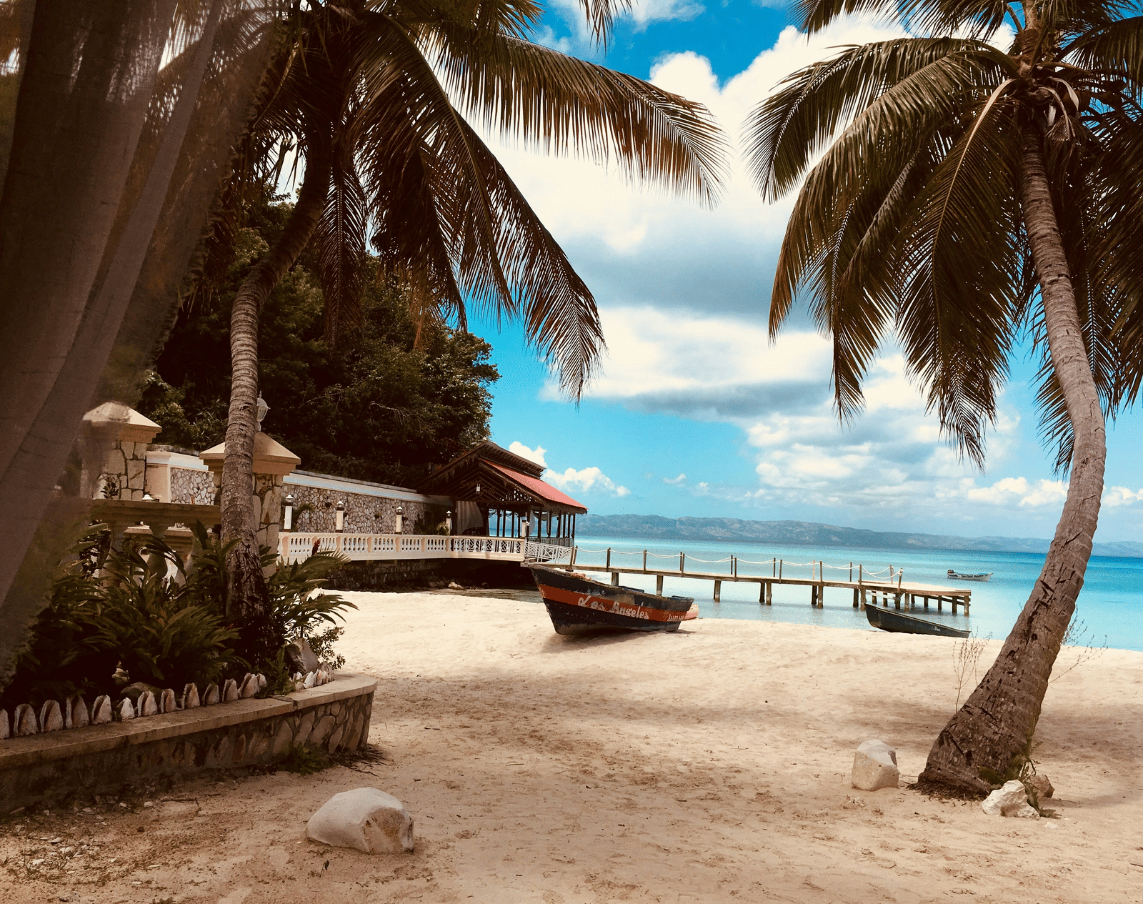 A tropical beach scene with white sand, palm trees, and a small wooden shack with a jetty stretching into the turquoise sea.