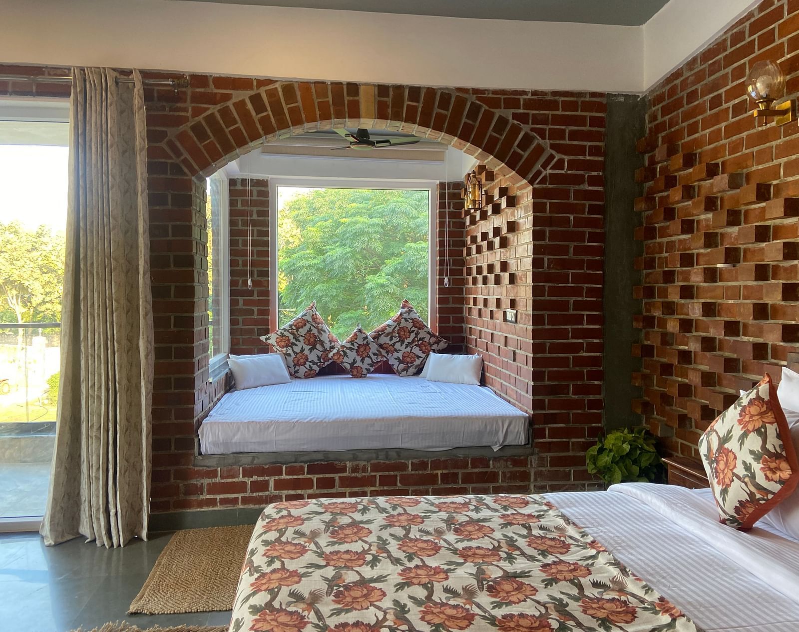 A bedroom at Tisya Farms, Gurgaon featuring a bed near a large arched window, brick walls, and a curtain on one side.
