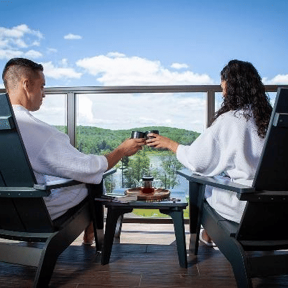 A couple in robes toasts with beverages while relaxing on a balcony with scenic views of green hills and a bright blue sky at YO1 Longevity & Health Resorts