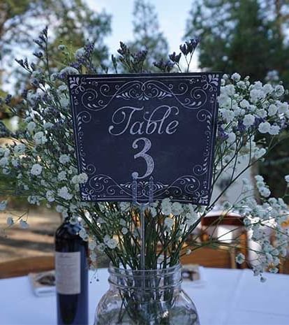 A wedding reception table is set outdoors with a 'Table 3' sign, floral arrangements, and a bottle of wine