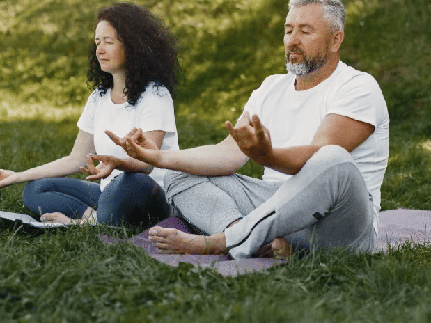 A middle-aged couple sits in a meditation pose on a yoga mat in a grassy park, with eyes closed, finding peace at YO1 Longevity & Health Resorts