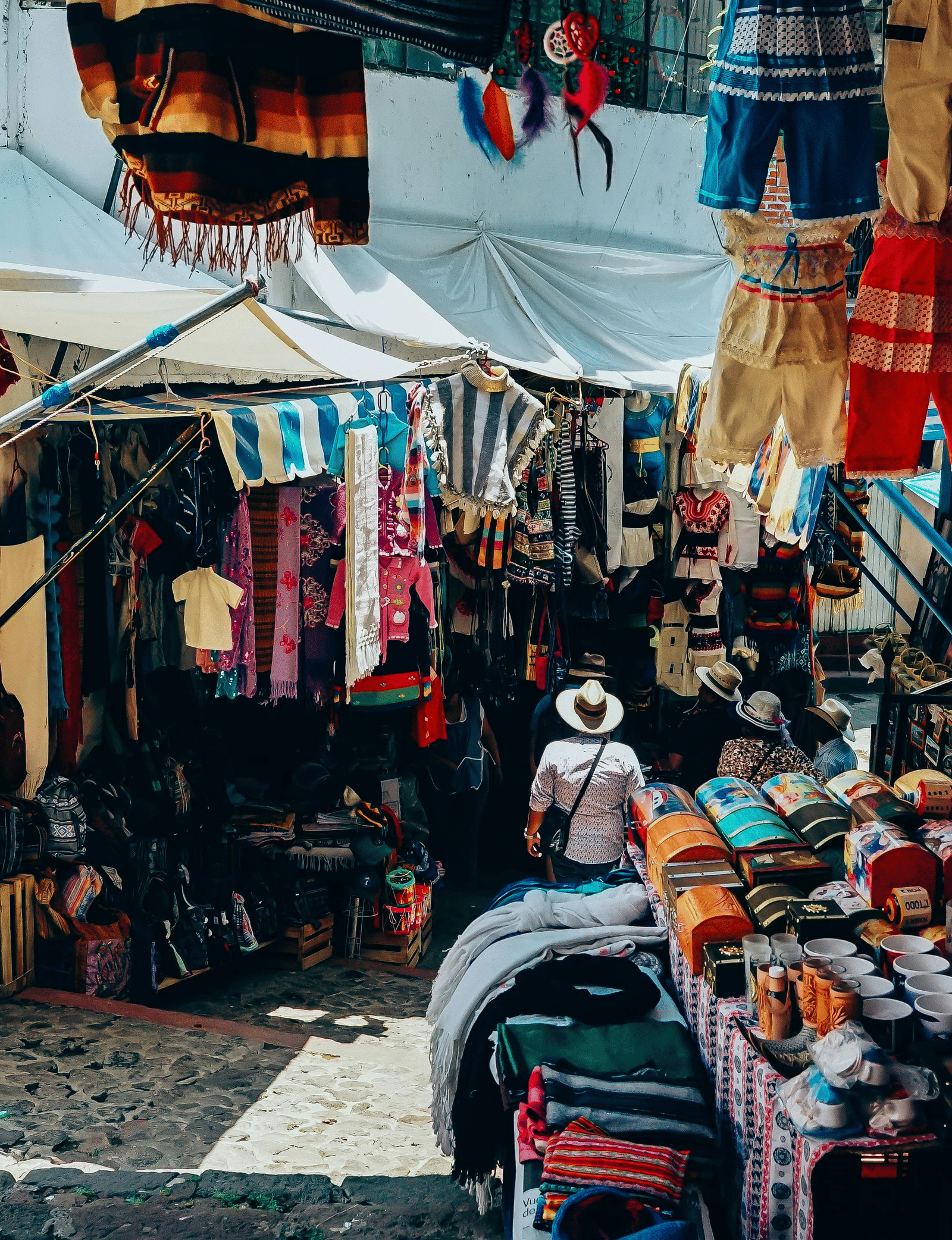 A bustling outdoor market stall selling textiles, clothing, and crafts, with a bright white awning overhead.
