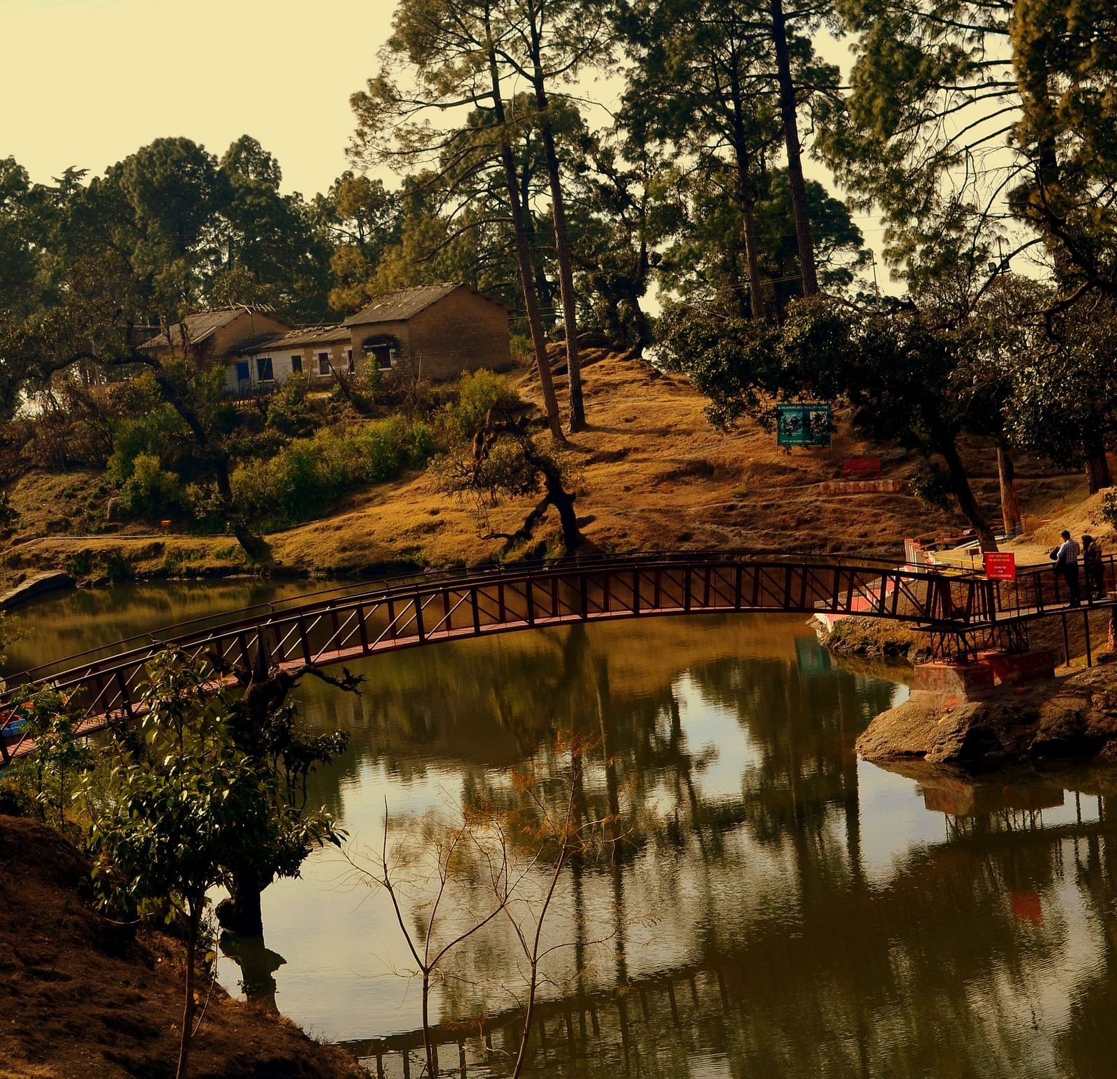 A bridge in the middle of a river, with a house and a shed for seating on the bank, surrounded by trees and two people standing on the bridge