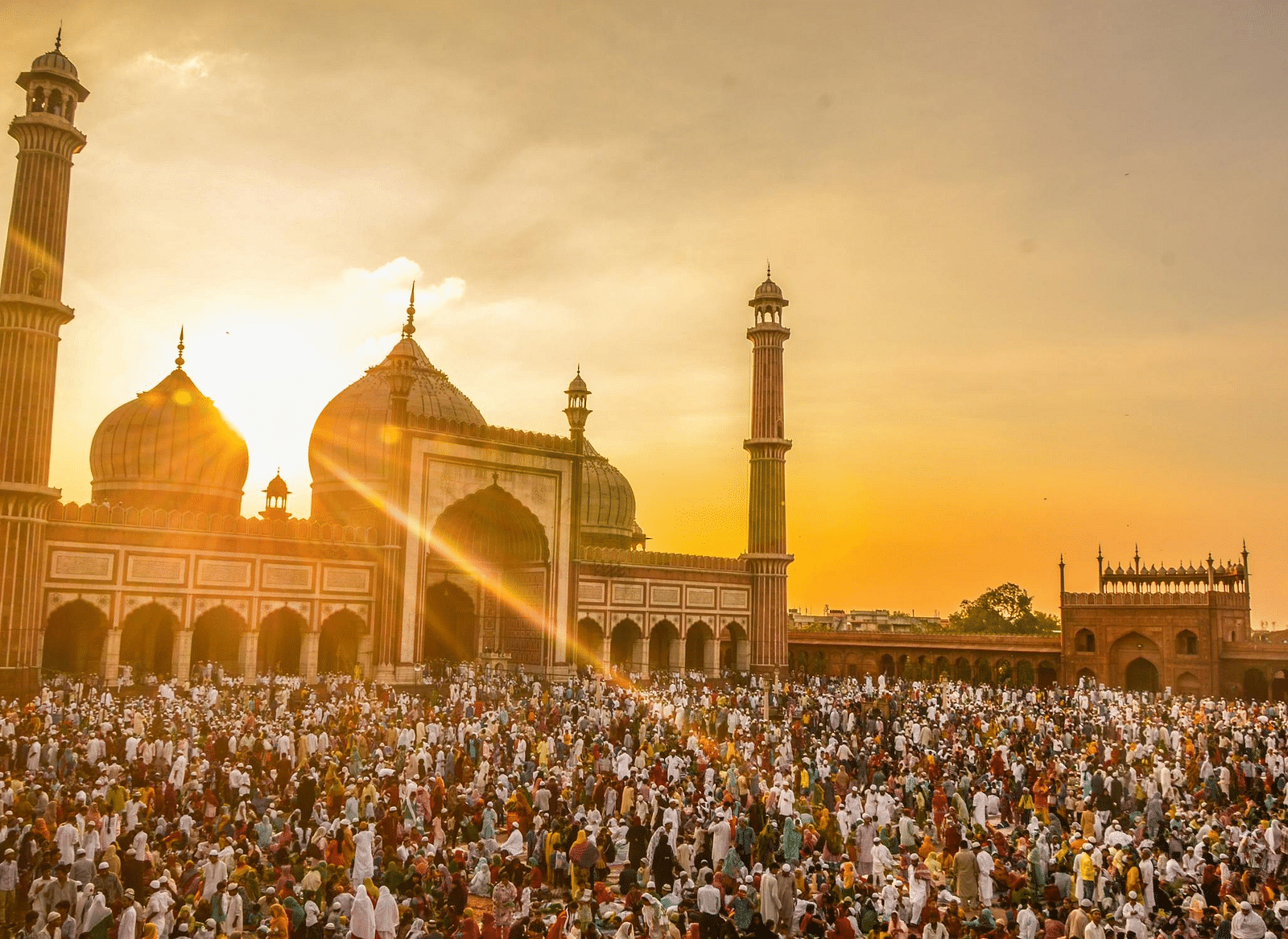 Image of a group of people gathering in front of a mosque with bright orange hued sky in the background.