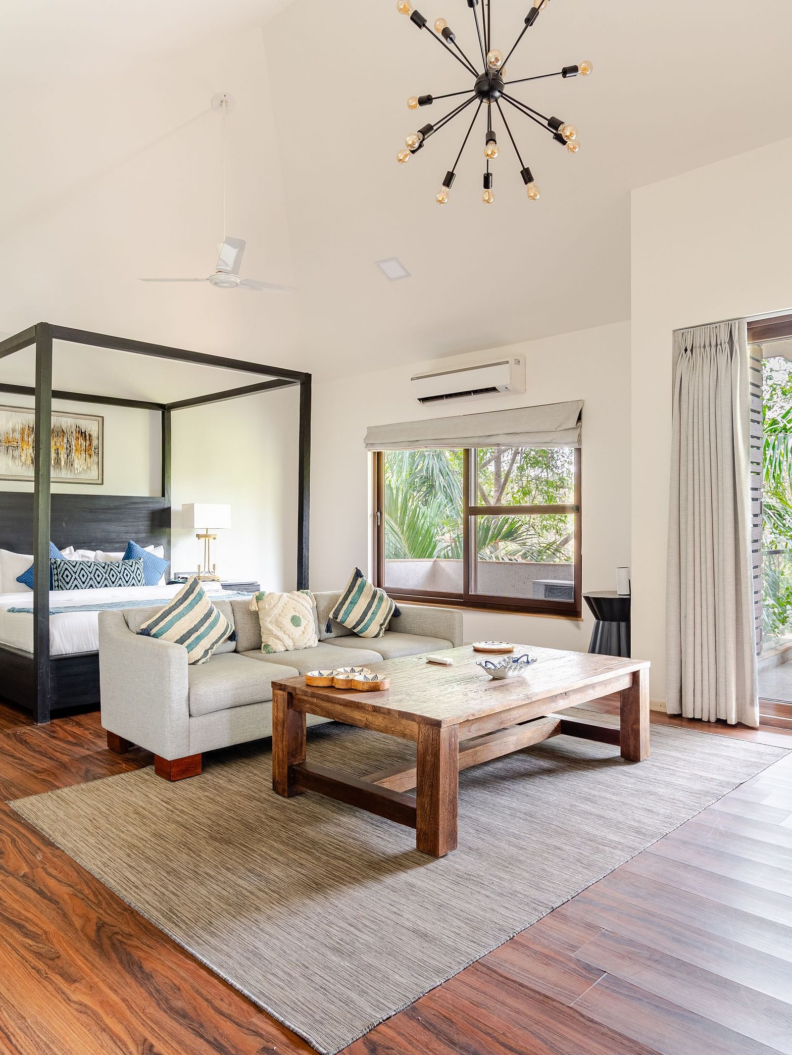Spacious, airy bedroom with high ceilings and warm wood flooring, featuring a four-poster bed with patterned pillows, a modern gray sectional sofa, a rustic wooden coffee table atop a neutral rug, a writing desk with books and flowers, and large glass door