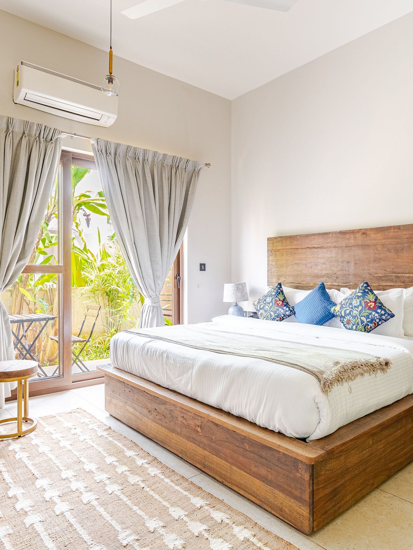 Bedroom at Lindsay Manor with a wooden bed, side drawer with a lamp, curtains, air conditioner, armchair, small coffee table, and a large window overlooking greenery.