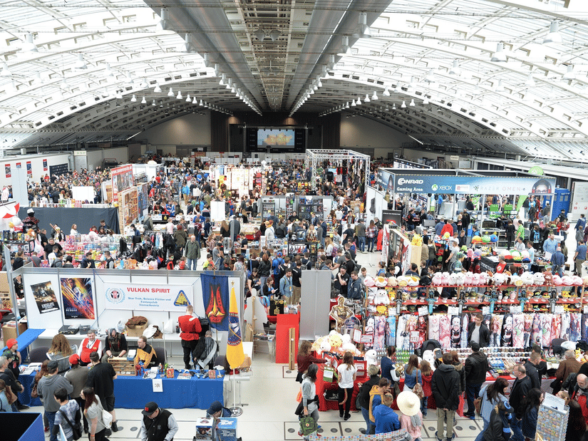 A bustling exhibition hall with a high vaulted ceiling, filled with many stalls and crowds of people at an event.