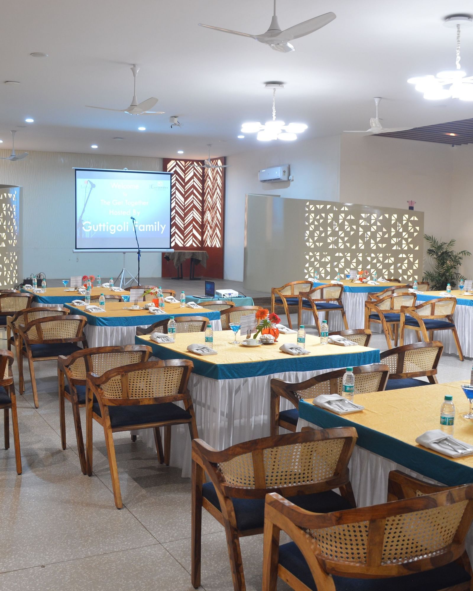 A banquet hall with a projector screen in the front with multiple tables and chairs under bright lights - Lords Eco Inn Belagavi