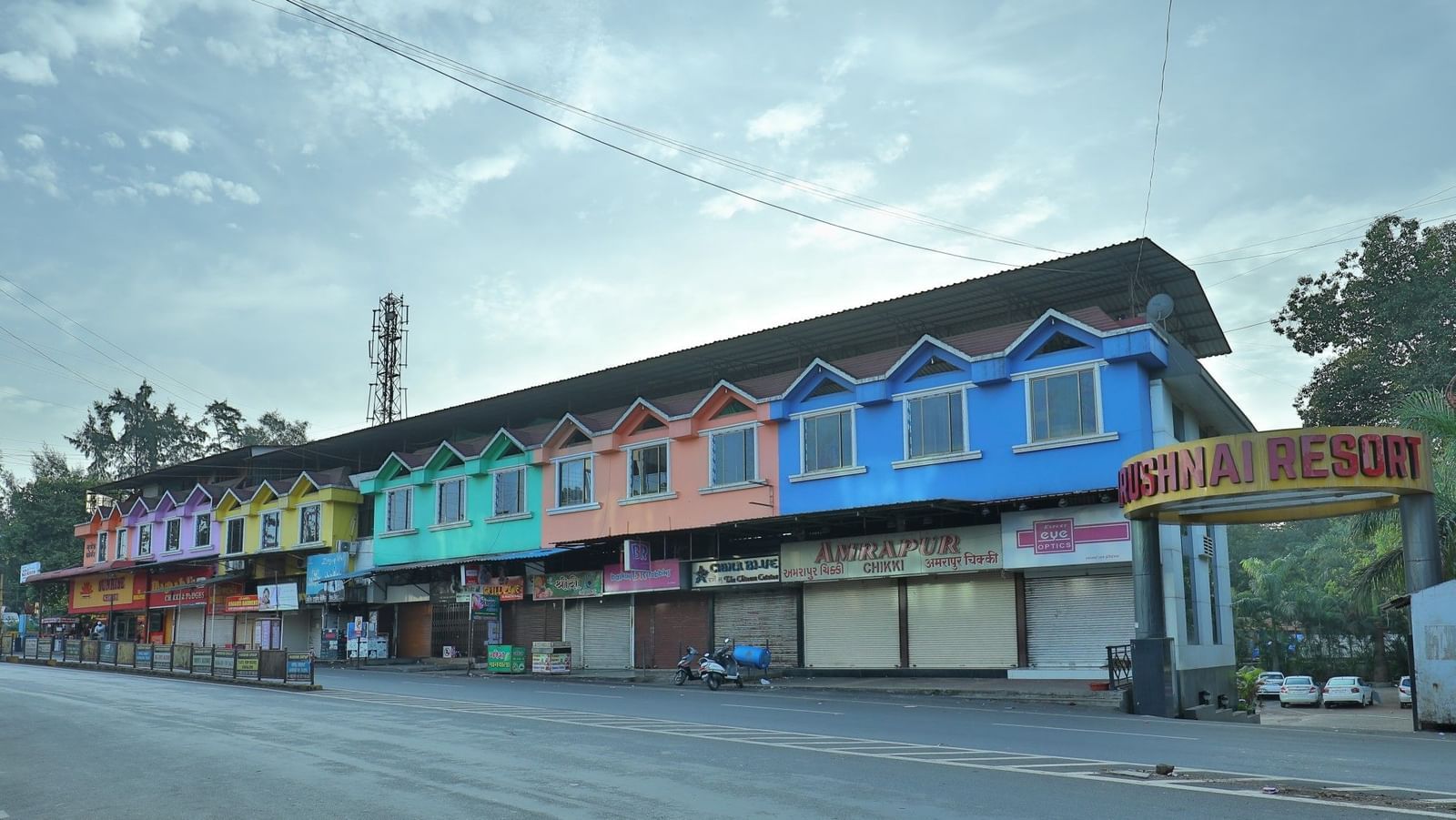 The Krushnai Resort sign marks a long building with colourful facade lining a quiet road under a cloudy sky.