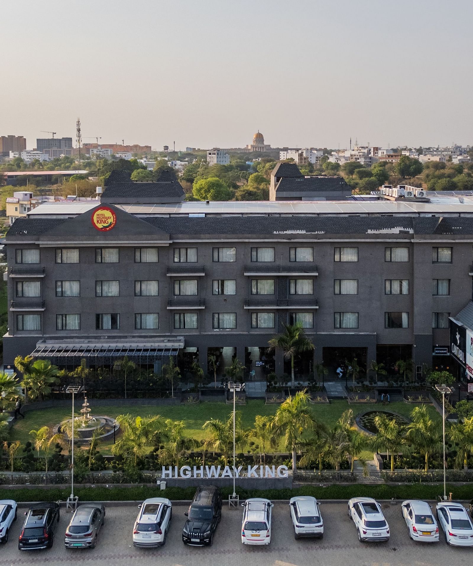 Aerial view of Hotel Highway King, Bagru, with landscaped gardens, parking area, and surrounding buildings in the background.