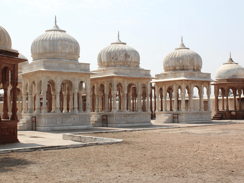 A row of ornate, white stone domed structures with arched pillars on a dusty ground under a clear sky.