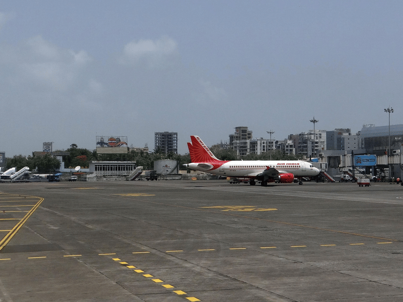 A white aeroplane with a red tail sits on a grey airport tarmac near city buildings under a bright sky.