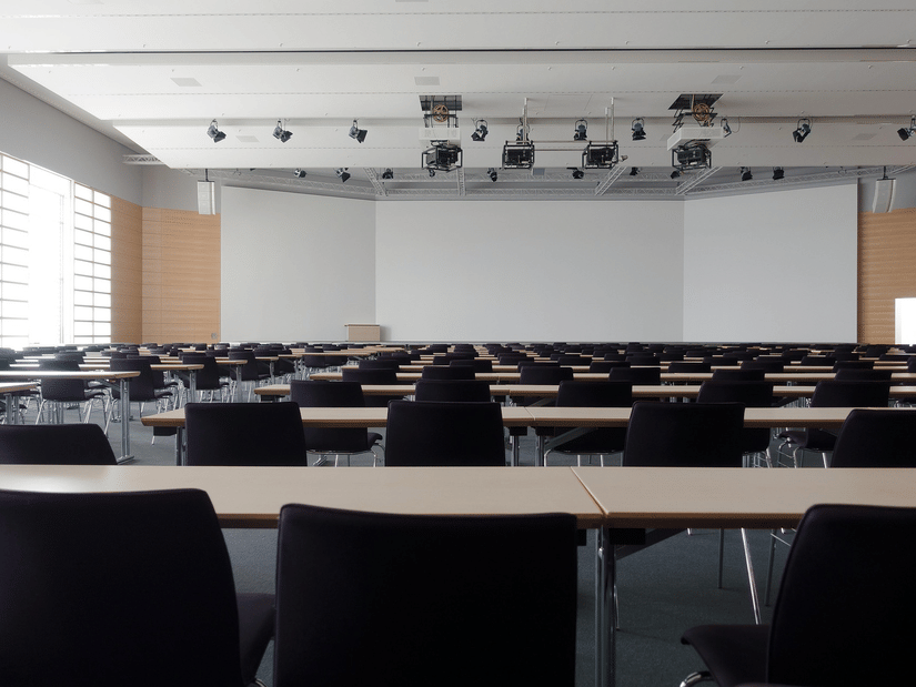 Rows of empty black chairs and wooden desks face a large white screen in a modern, brightly lit lecture theatre.