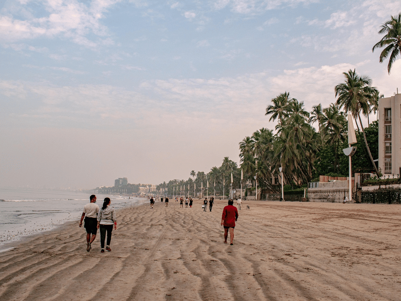 People walk along a wide, sandy beach lined with tall palm trees and buildings under a hazy, pale sky.