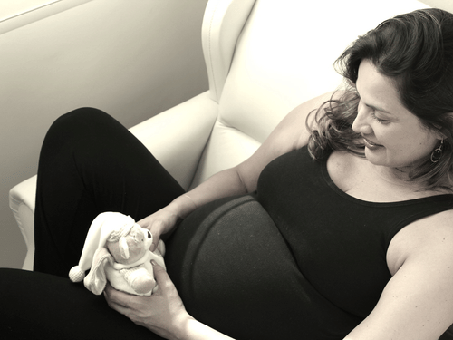 A smiling pregnant woman holds a stuffed bunny near her baby bump while sitting in a chair.