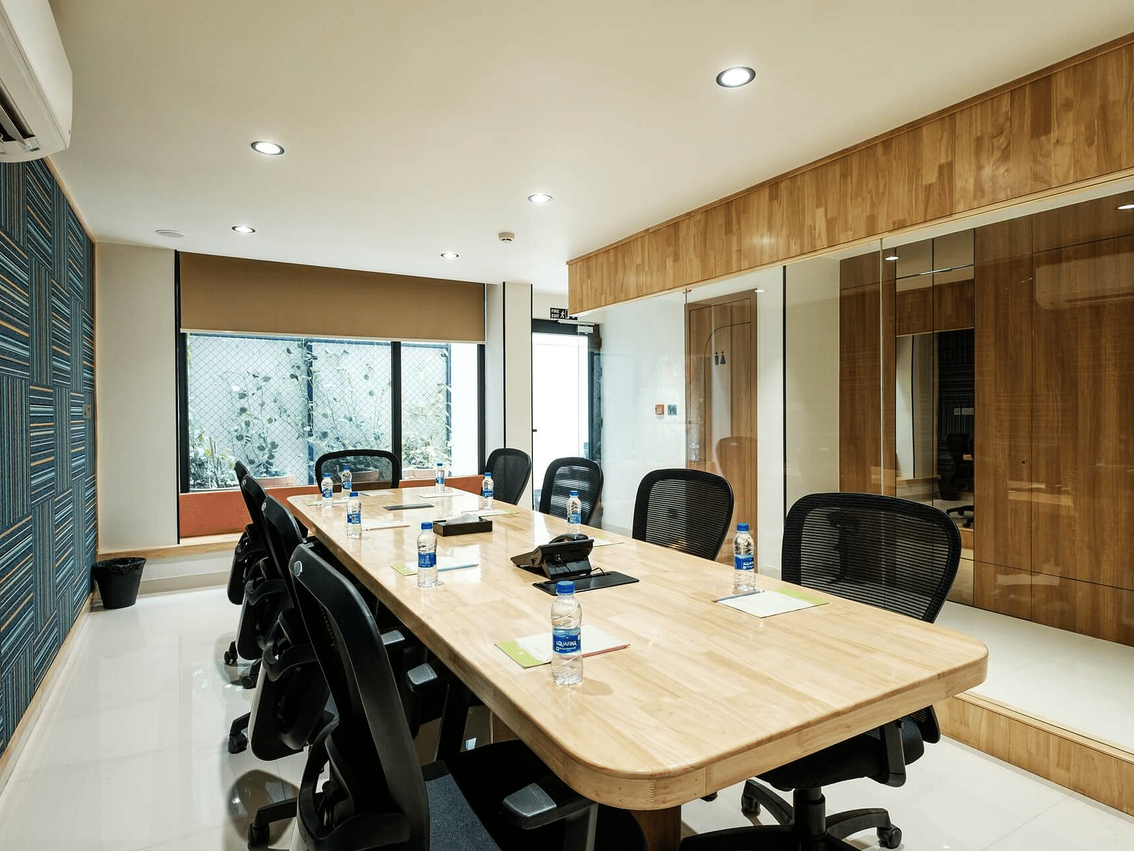 Diagonal view of the full boardroom at Indie Stays, Jaipur, with chairs arranged along the sides, a wall-mounted AC, and large windows at the end.