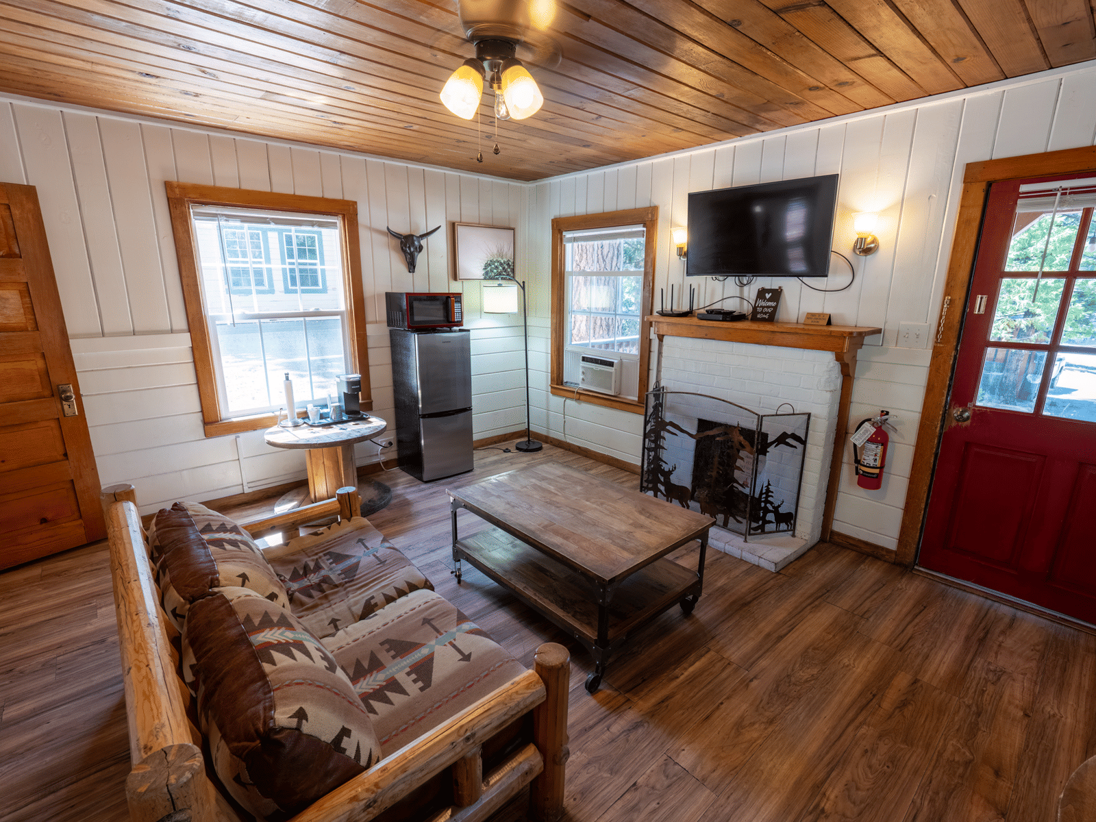 A cabin living area with wood-paneled walls and ceiling, rustic wooden furniture, a fireplace, and a wall-mounted TV - Shaver Lake Village Hotel
