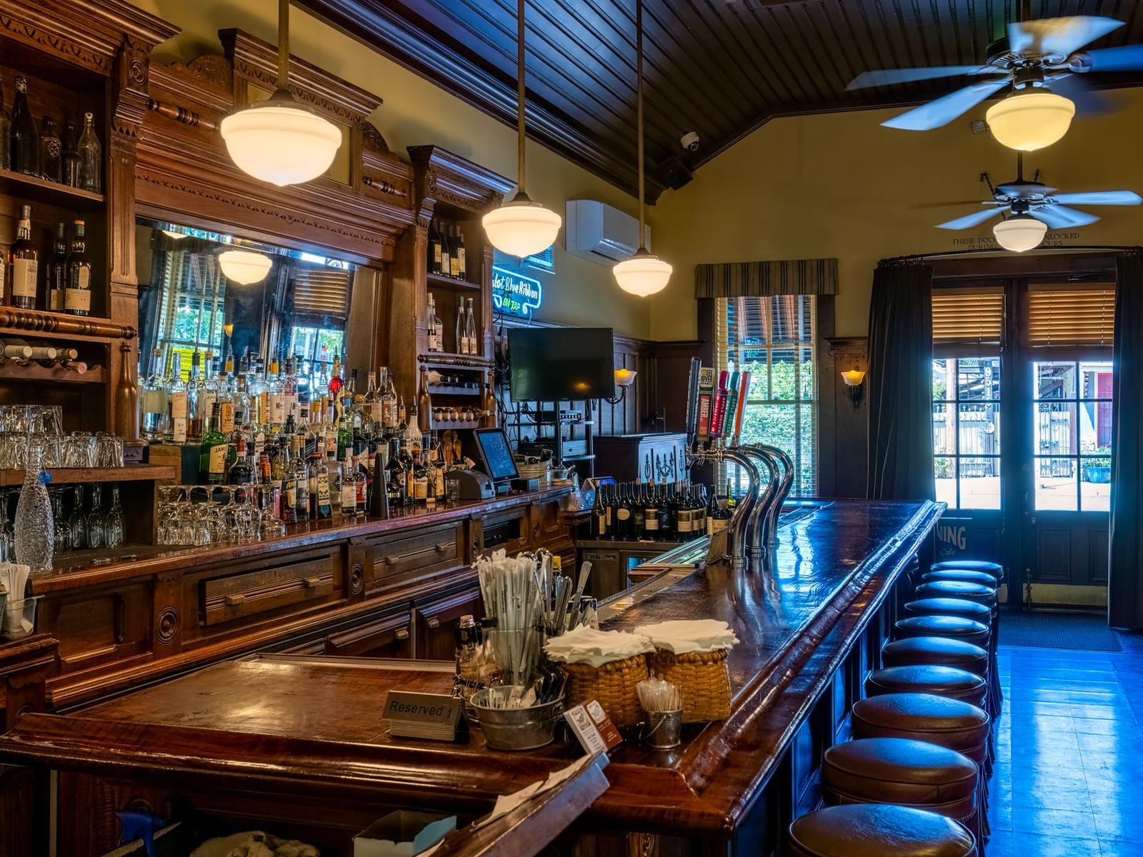The rustic interior of a lively bar with a long wooden counter, bar stools, and various beverages at The Tallman Hotel.