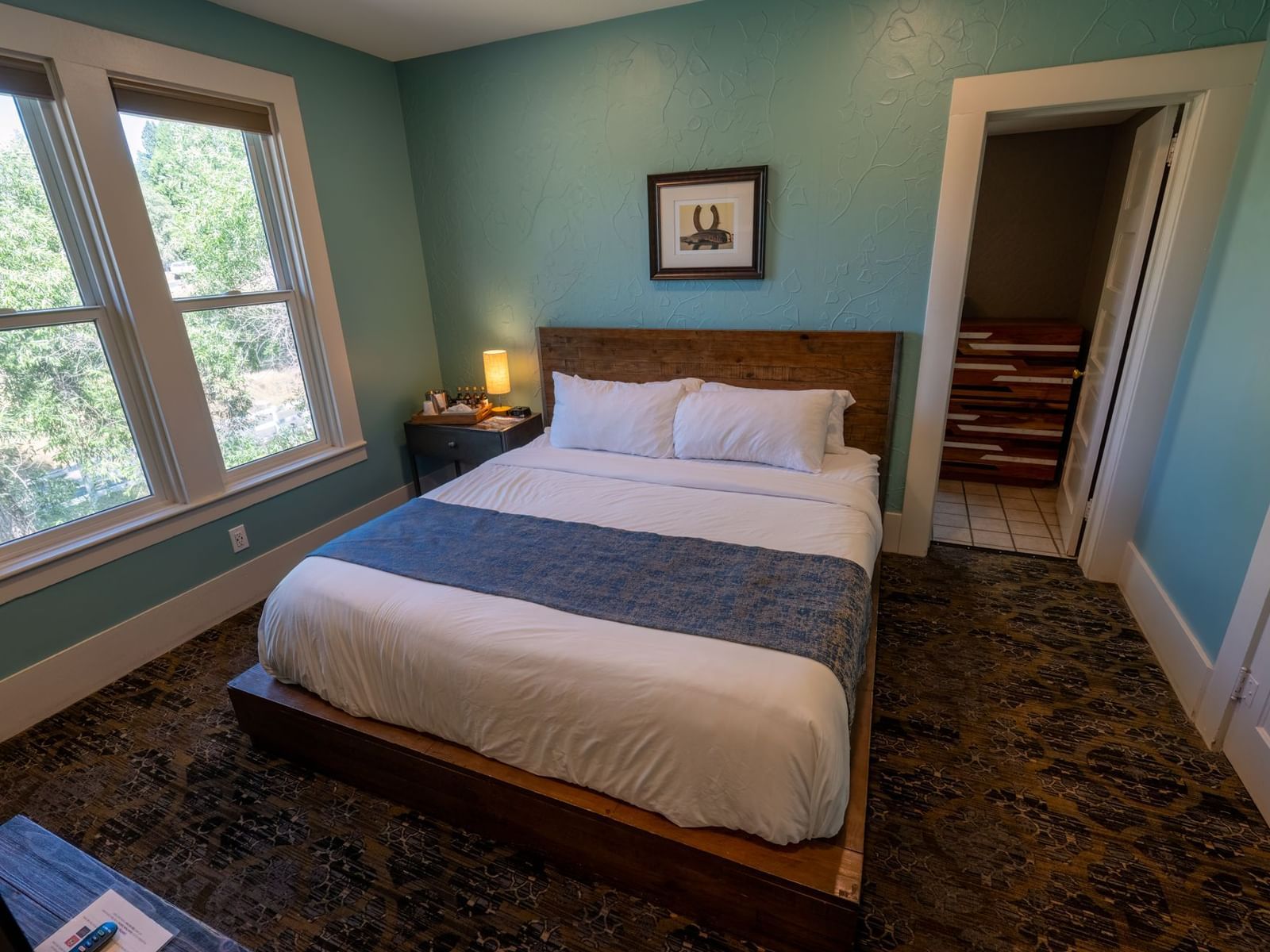 Corner view of a Family Suite at The Groveland Hotel featuring a plush bed, carpeted flooring, and a wood-framed glass window offering a view of the greenery nearby.