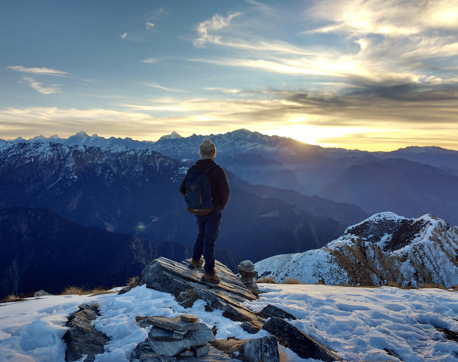 A traveller standing on a snowy mountain ridge overlooking dramatic Himalayan ranges at sunrise or sunset.