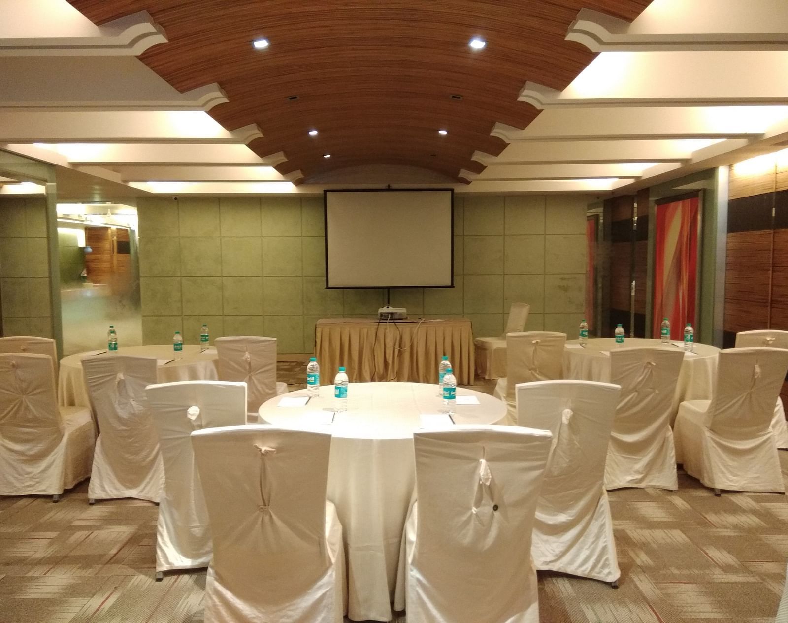 Banquet hall at Mirage Hotel, Mumbai featuring ample seating area, round tables covered in white sheets and carpet flooring.