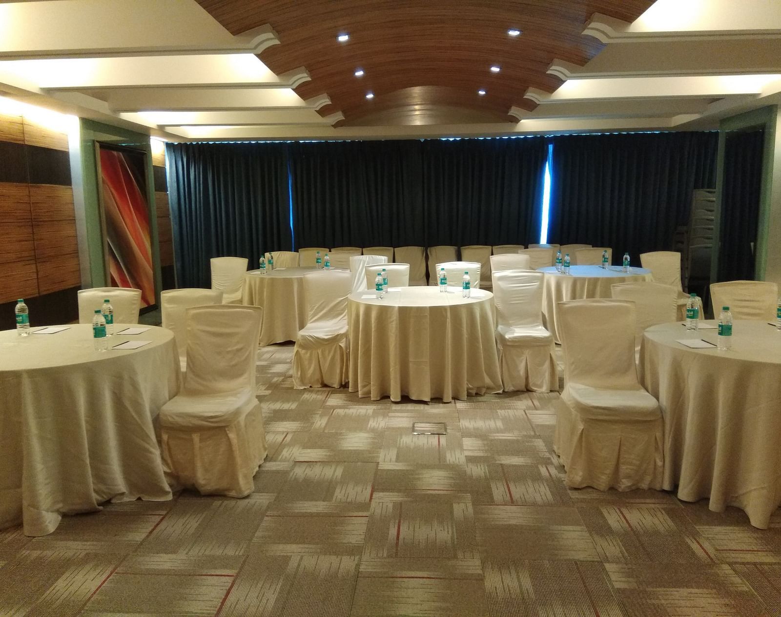 Banquet hall at Mirage Hotel, Mumbai featuring ample seating area, round tables covered in white silk covers and carpet flooring.