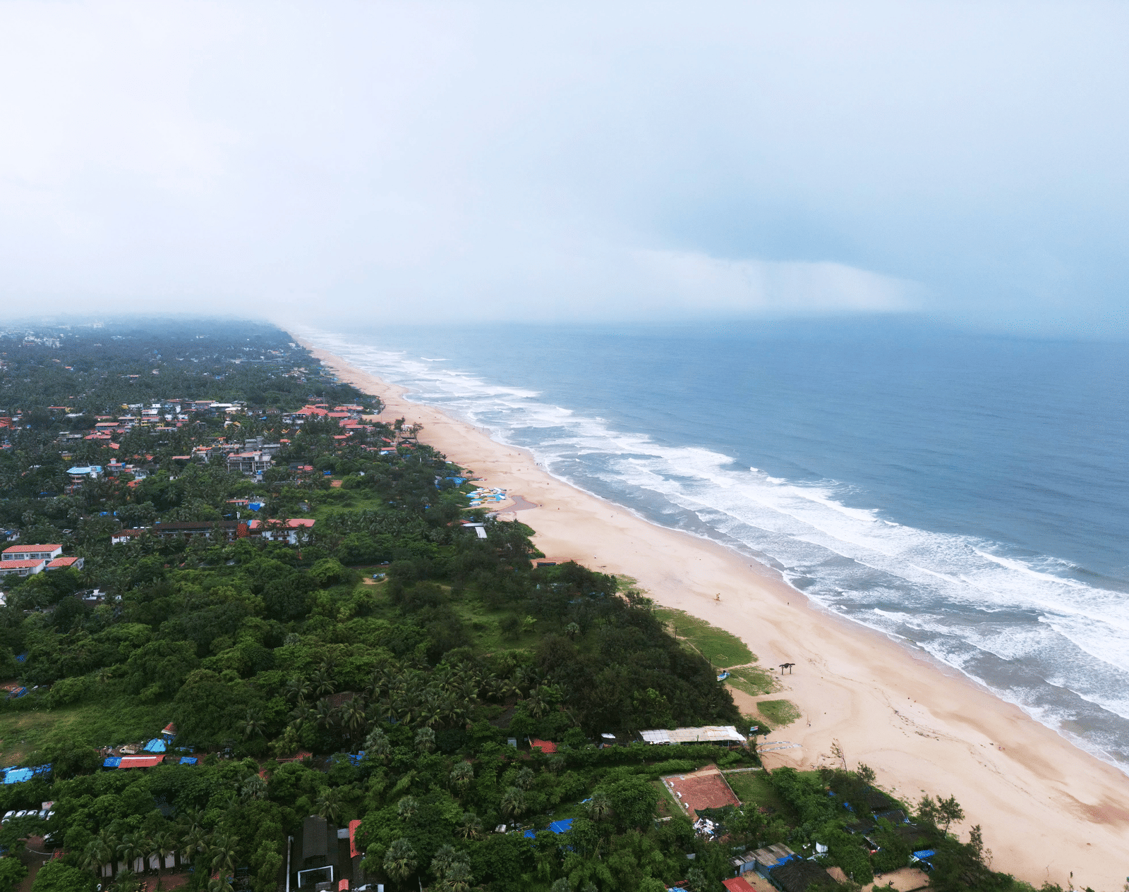 A aerial view of a long, curving beach, where waves meet sand and dense green settlements line the coast under a cloudy sky.