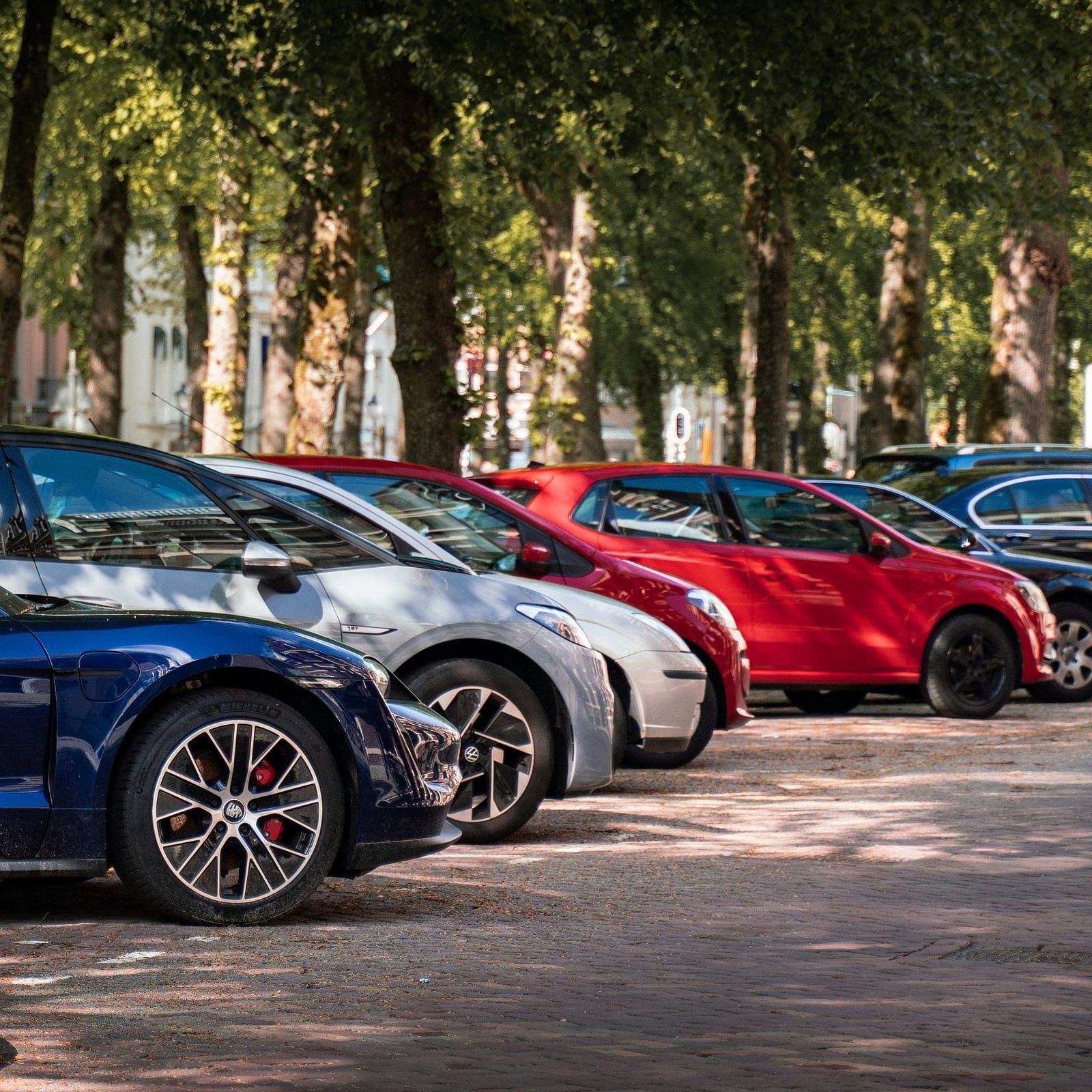 A row of parked cars on a tree-lined street, with varying colours and models on a cobblestone pavement