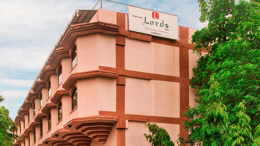 The facade of Lords Inn Vadodara featuring a red and pink toned building with the hotel signboard and lush greenery under partly clouded sky.